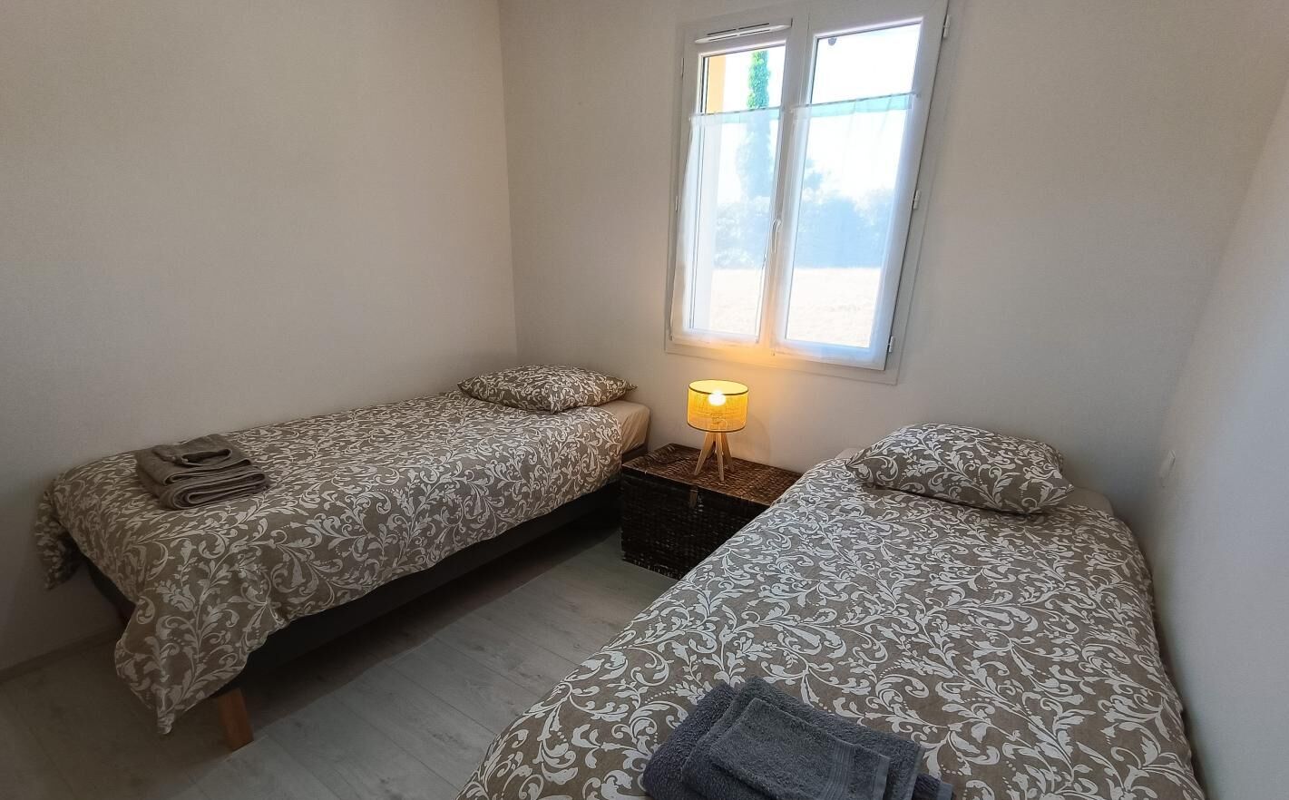 A small, well-lit room with two single beds, a nightstand with a lamp, and a window providing natural light.