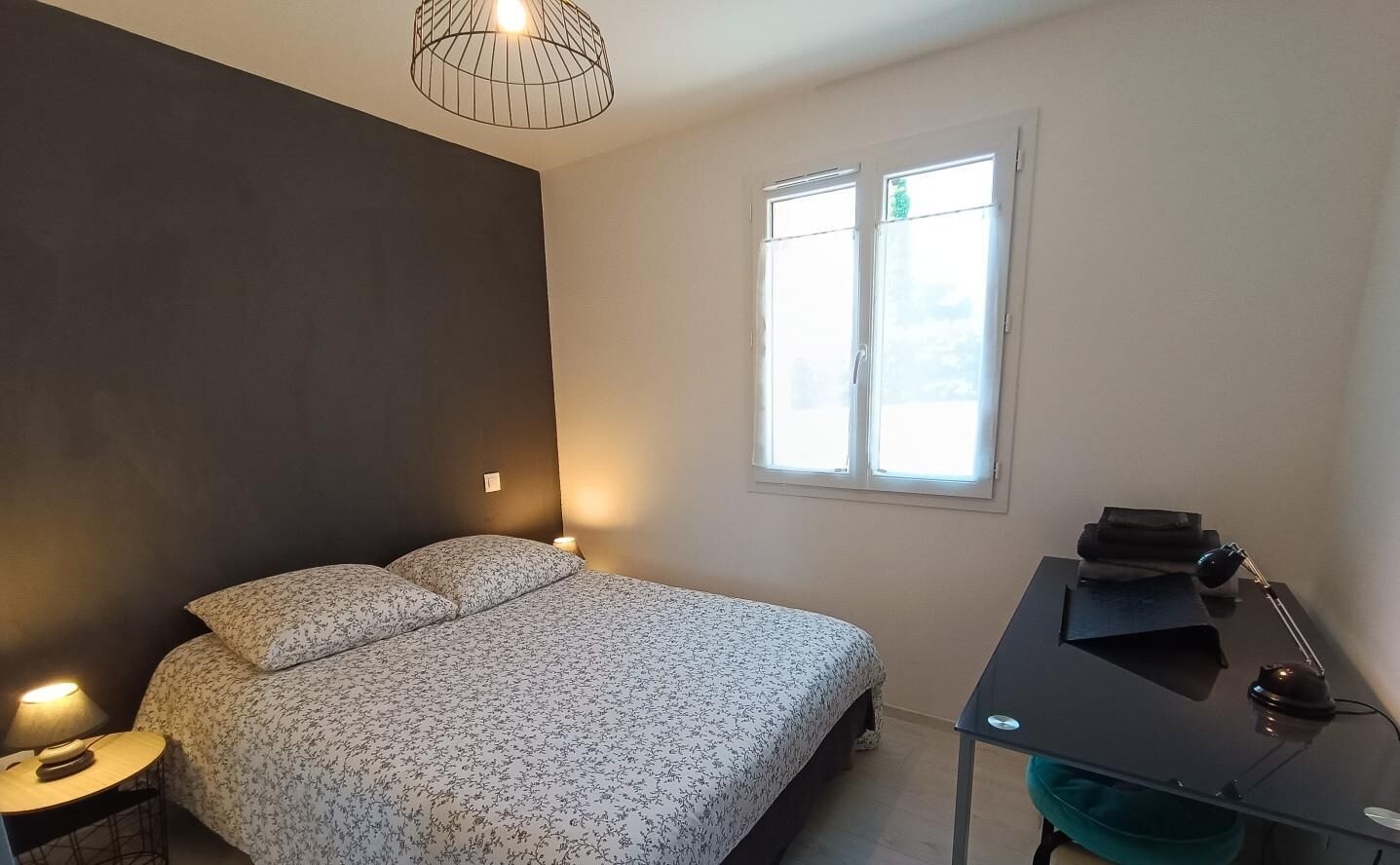 Quarto com cama, mesa de cabeceira, luminária, escrivaninha, cadeira e janela.