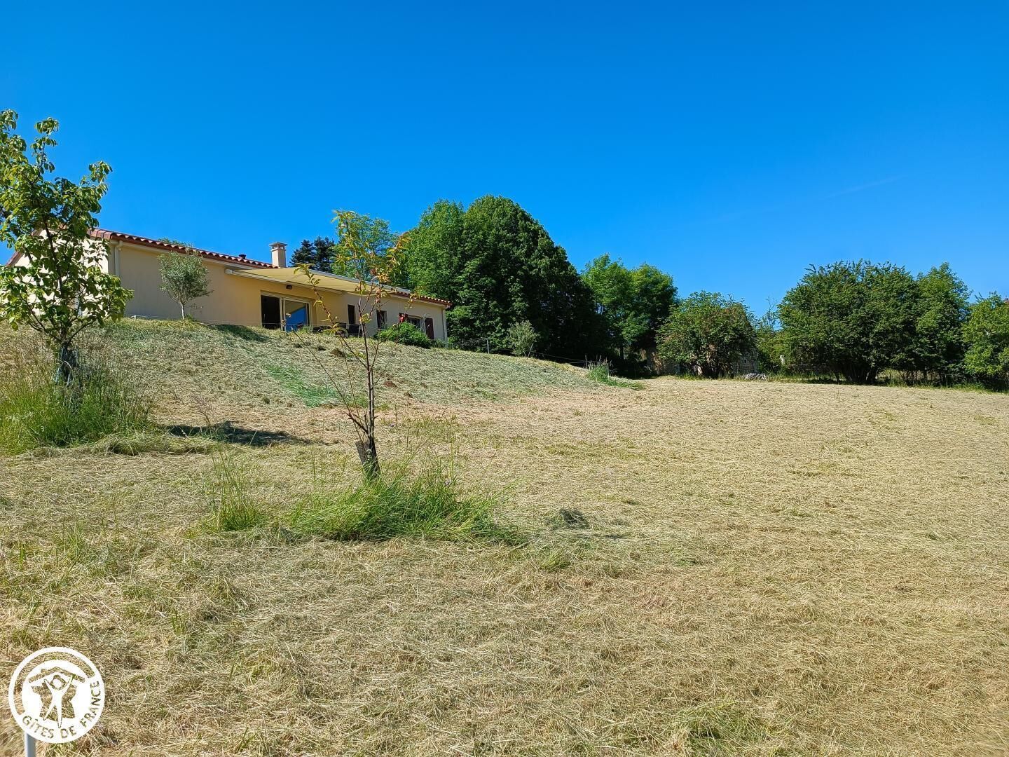 A house with a red-tiled roof and a yellow facade, surrounded by a grassy field and trees.