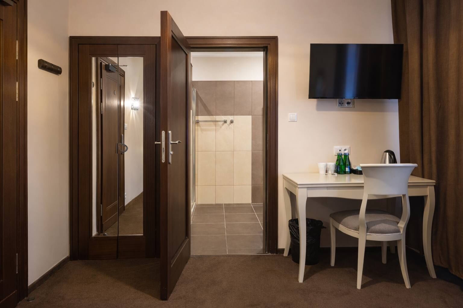 A hotel room with a wooden door, a shower area, a desk with a chair, a flat-screen TV, and a small table with bottles and cups.
