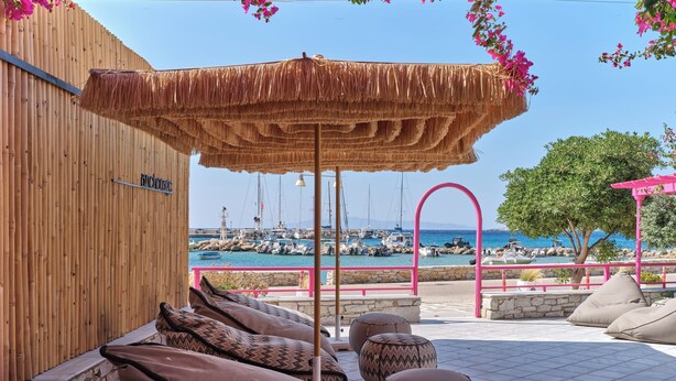 A beachside seating area with a thatched umbrella, stone walls, and a view of boats at a marina. - Anchorage Paros  (Piso Livadi)