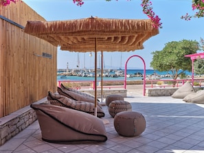 Un área de descanso frente a la playa, con una sombrilla de paja, muros de piedra y vistas a barcos en un puerto deportivo.