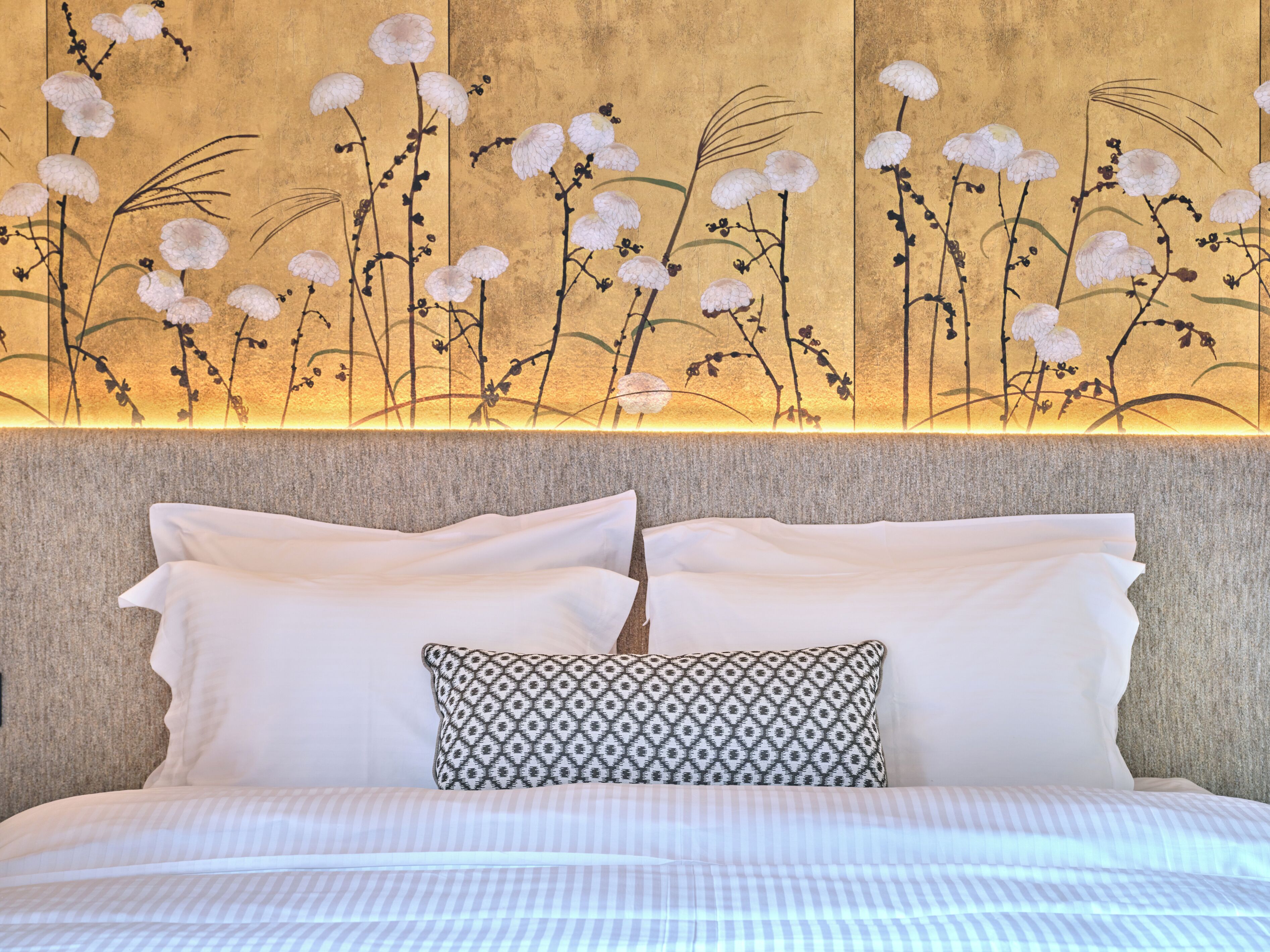 Una cama con sábanas blancas y un cojín estampado, frente a una pared con un mural floral.