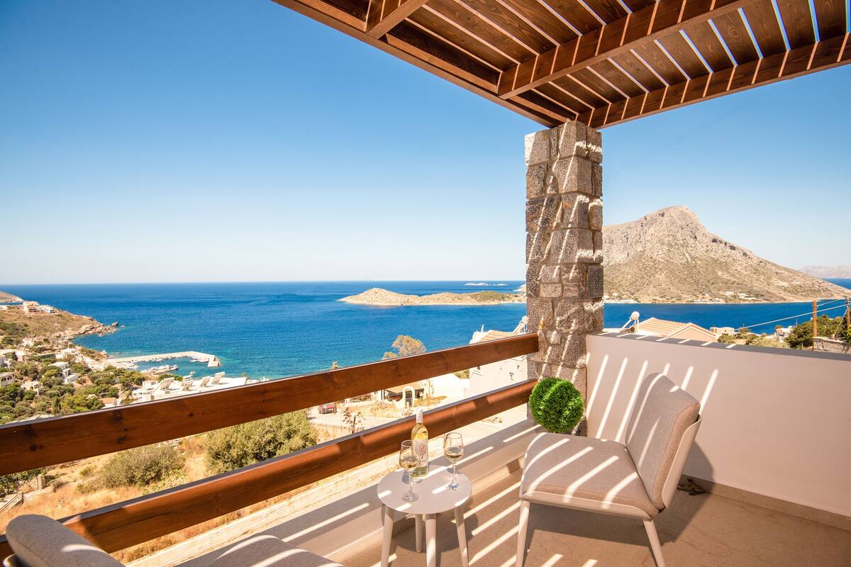 A balcony with a view of the sea, a stone pillar, and a table with glasses and a bottle.