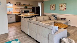 A modern living room with a sectional sofa, a wooden coffee table, and a small round table. There is a kitchen area with a refrigerator, microwave, and various kitchen items on the counter. The room has light green walls and a ceiling fan. - Pet-Friendly Gulf Front Escape nestled in the Dunes (Dauphin Island)