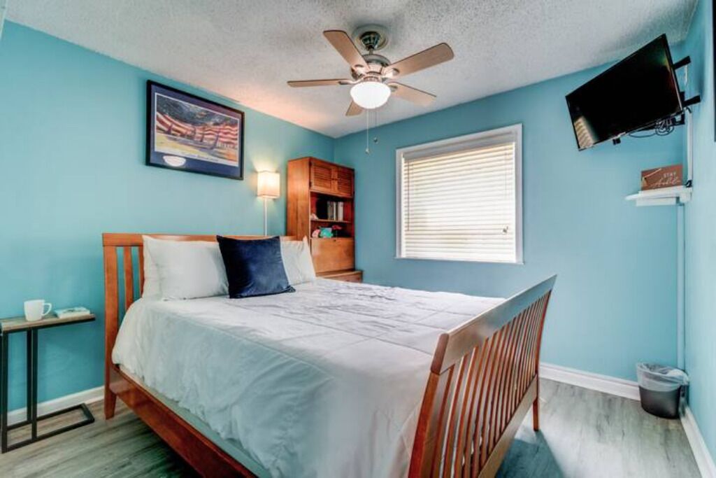 A bedroom with a bed, a ceiling fan, a television mounted on the wall, a window with blinds, and a bookshelf.