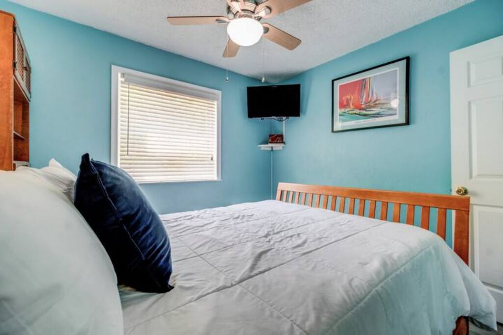 Une chambre à coucher avec un lit, un ventilateur de plafond, une fenêtre avec des stores, un téléviseur fixé au mur et un tableau encadré accroché au mur.