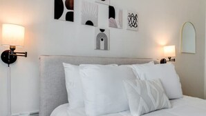 Ein ordentlich bezogenes Bett mit weißen Leinen und Kissen, ein Nachttisch mit einer weißen Tasse sowie ein an der Wand befestigtes Kunstwerk.