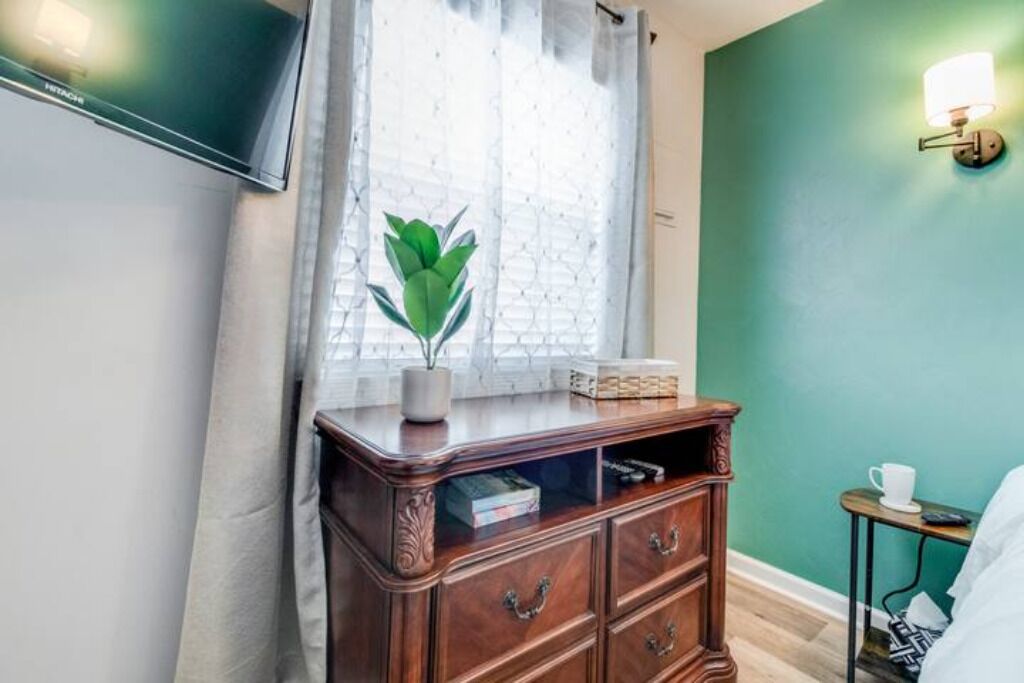 A room with a wooden dresser, a potted plant, a window with sheer curtains, a wall-mounted light fixture, and a bed with a white comforter.
