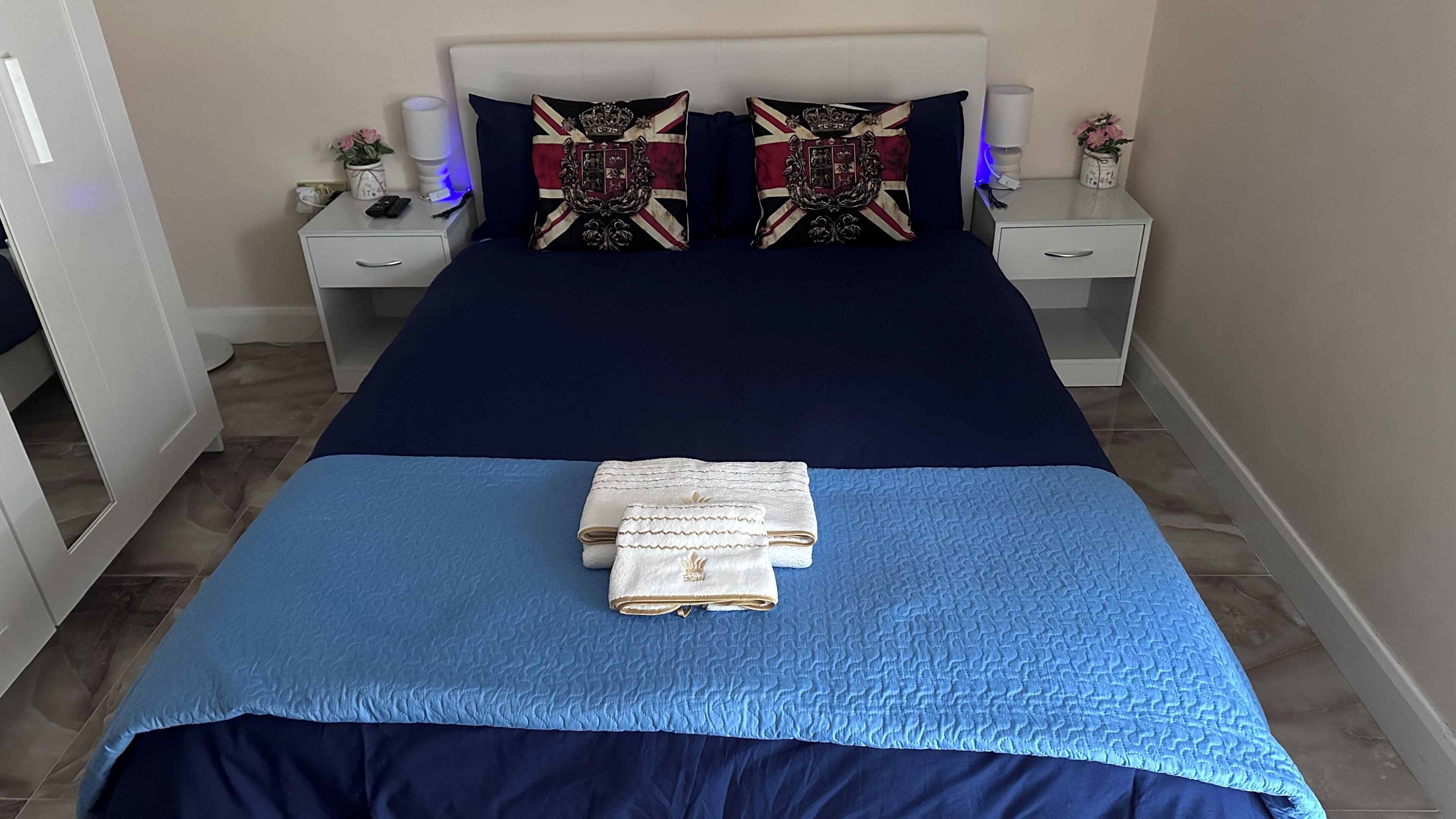 A neatly made bed with a blue and navy blue bedspread, two decorative pillows, a folded towel, and a bedside table with a lamp and a vase of flowers.