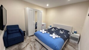 A bedroom with a bed, a blue armchair, a wardrobe, a bedside table, and a nightstand. - Royal Modern Studio Heathrow London (Feltham)