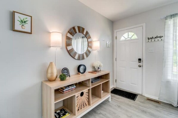 Un couloir avec une console en bois, un miroir rond, des lampes fixées au mur et une porte sur laquelle est affiché un panneau indiquant : « Veuillez déposer les clés ici ».
