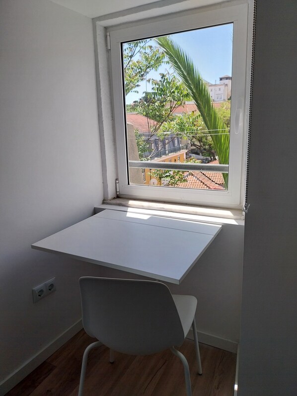 Une pièce avec un bureau et une chaise blancs, une fenêtre donnant sur des immeubles et des arbres.