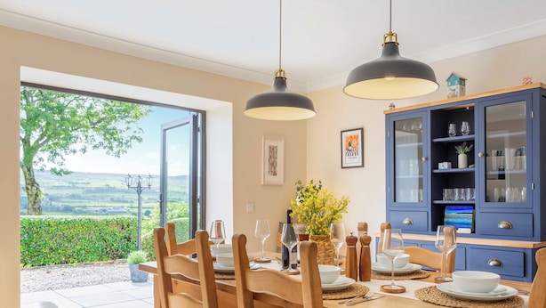 Un comedor con una mesa dispuesta para cuatro personas, sillas de madera, un armario con puertas de vidrio y vistas a través de un ventanal.