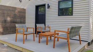 A deck with four wooden chairs and a table, a black door, and a window on a house with siding. - Serene 2-bedroom apartment with WiFi in Ellsworth with a backyard stream. (Ellsworth)