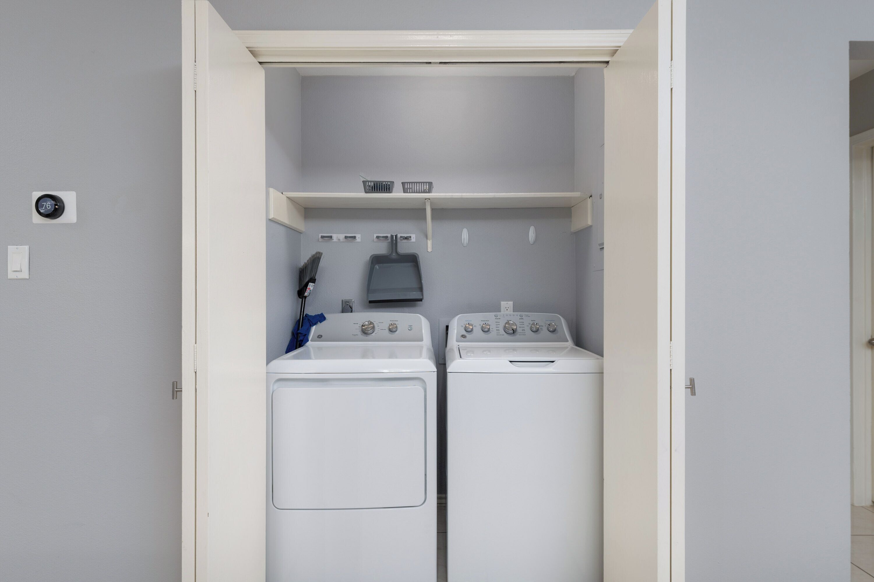 A laundry room with two stacked washers and dryers, a shelf above, and a thermostat on the wall.