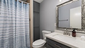 A bathroom with a shower curtain, toilet, sink, mirror, and a soap dispenser. - The 3 Queen Suite by 802 & Paredes (Brownsville)