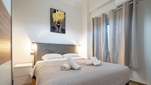 Cama bem arrumada com colcha acolchoada e duas toalhas enroladas, mesa de cabeceira com abajur e uma obra de arte pendurada na parede.