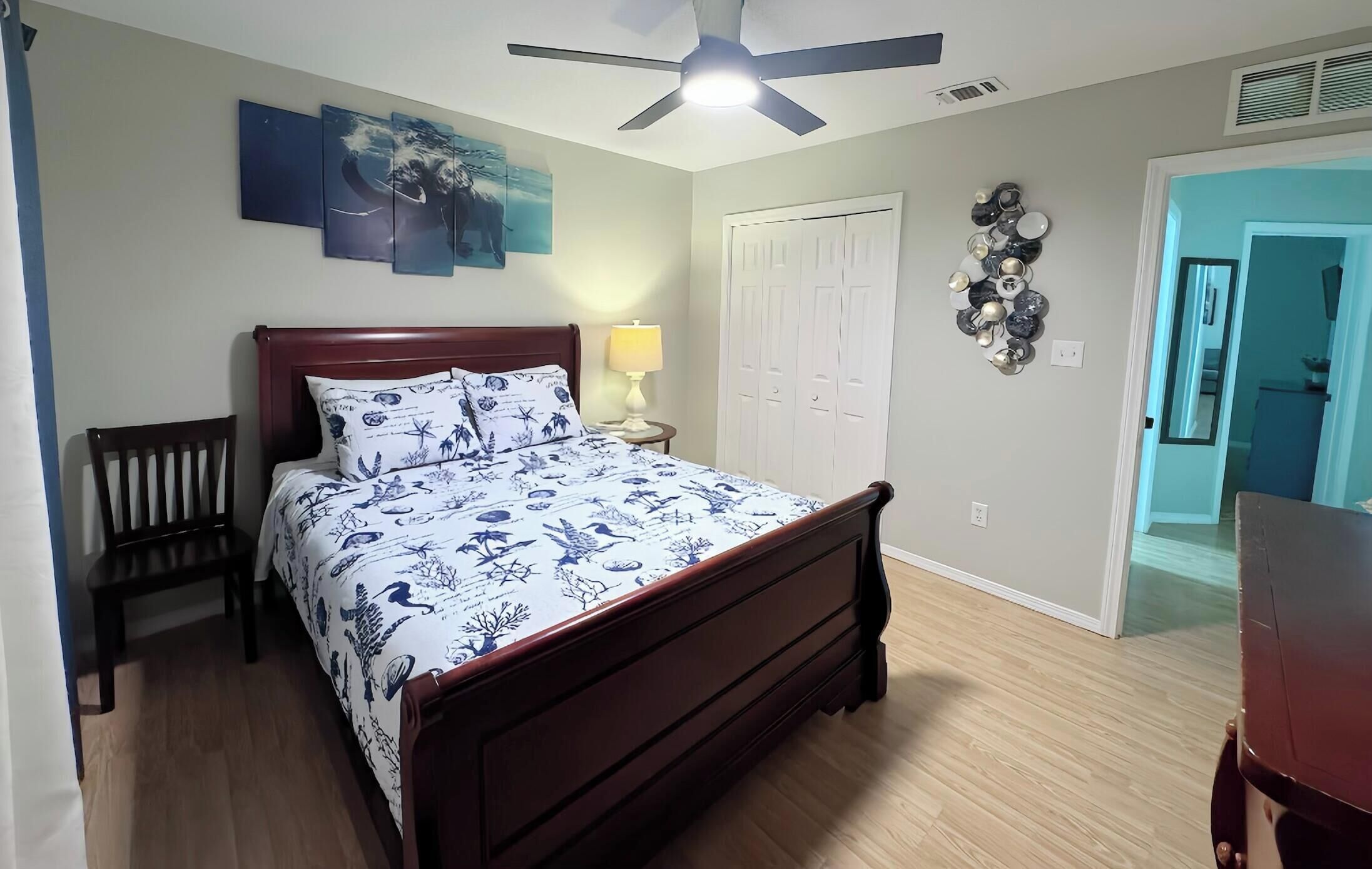 A bedroom with a bed, a chair, a ceiling fan, a wall-mounted lamp, a door, and a wall decoration.