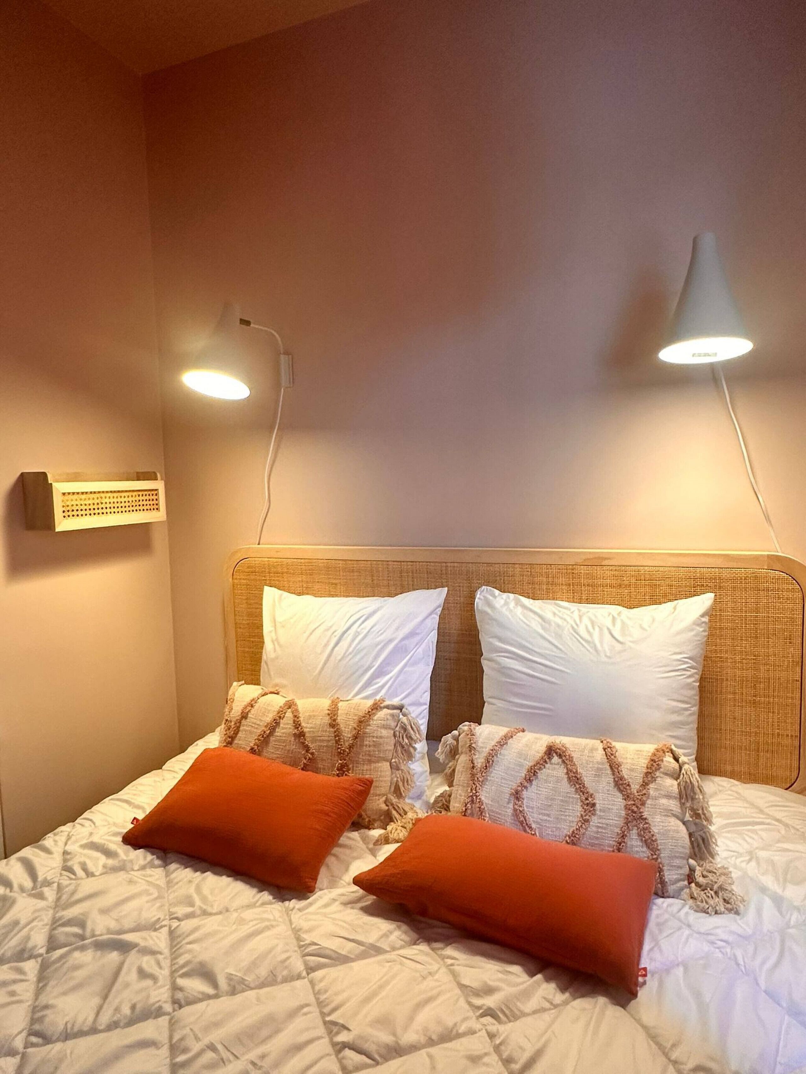 Ein Bett mit weißer Bettwäsche und zwei orangefarbenen Kissen, ein Kopfteil und zwei an der Wand befestigte Lampen.