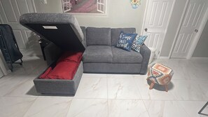 A grey sofa with a red cushion, a blue pillow with 'home sweet home' text, a small patterned stool, and a window with a pink floral view.