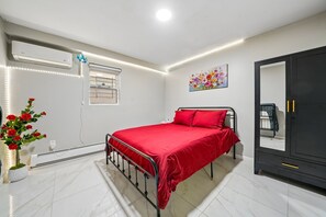 Un dormitorio con cama, cómoda, radiador, ventana con vistas y aire acondicionado instalado en la pared.