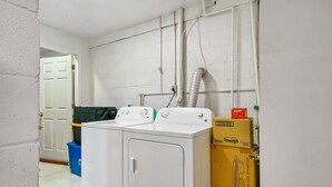 Una sala de bugaderia amb una rentadora i assecadora de càrrega frontal, un cubell de brossa blau i una caixa per a una barbacoa elèctrica amb una olla calenta.
