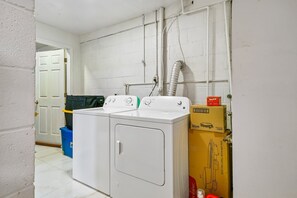 Une buanderie équipée d’un lave-linge et d’un sèche-linge à chargement frontal, d’une poubelle bleue et d’une boîte contenant un appareil de cuisson électrique avec une plaque chauffante.