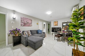 Un salon moderne avec un canapé d’angle gris, un coin repas comprenant une table et des chaises, et une grande plante verte.