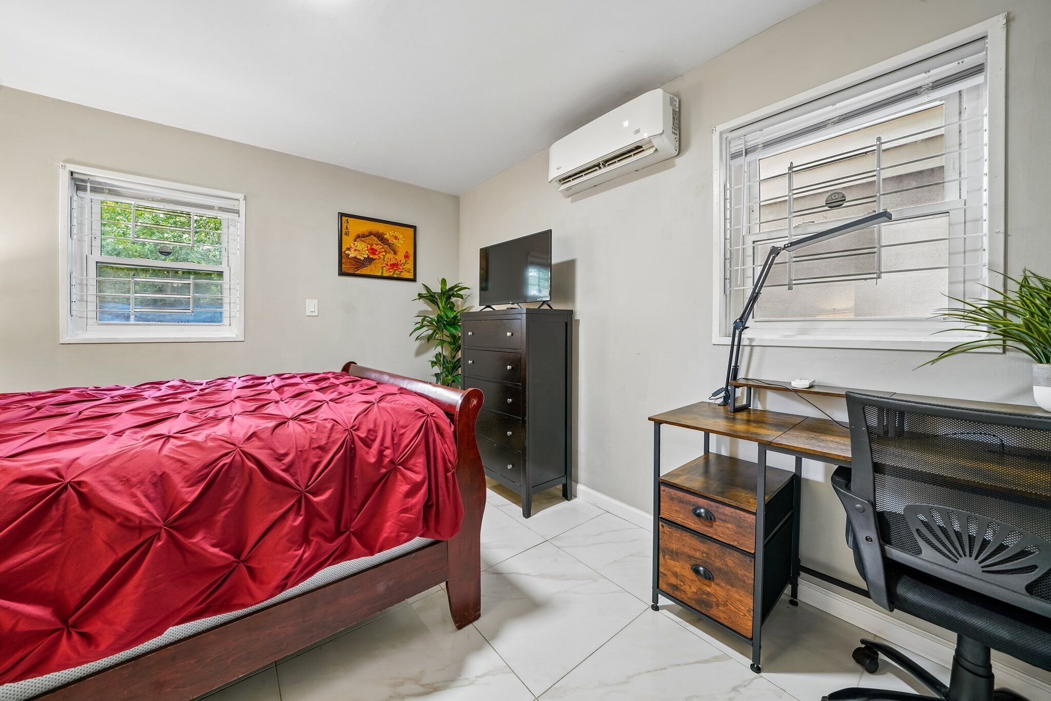 Une chambre à coucher comprenant un lit, un bureau avec une chaise, une commode et deux fenêtres équipées de stores.