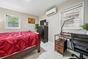 Une chambre à coucher comprenant un lit, un bureau avec une chaise, une commode et deux fenêtres équipées de stores.