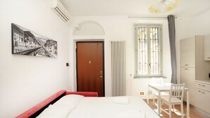 A hotel room with a bed, a desk, a chair, a radiator, a window with curtains, a door, and a wall-mounted air conditioner. - Moro 137 Apartment Navigli  (MILANO)