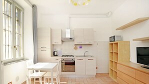 A compact kitchen with white cabinets, a gas stove, a sink, and a microwave. There is a dining table with chairs, and a TV on a wooden stand. - Moro 137 Apartment Navigli  (MILANO)