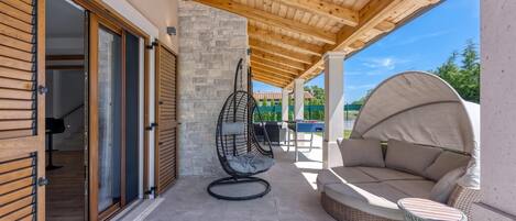 A covered outdoor patio with a wooden ceiling, a stone wall, a swing chair, a sofa, and a table.