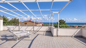 Une terrasse sur le toit, dotée d’une structure métallique, d’un sol carrelé et offrant une vue sur la mer et les bâtiments en arrière-plan.