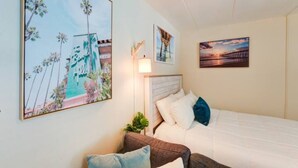 A hotel room with a bed, a nightstand, and a large painting of a coastal scene. - Sunny Days Beach Studio on the Boardwalk w/Pool (Virginia Beach)