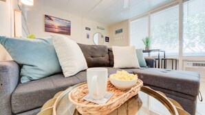 A modern living room with a grey sofa, a coffee table with a tray, and a bowl of cereal. - Sunny Days Beach Studio on the Boardwalk w/Pool (Virginia Beach)