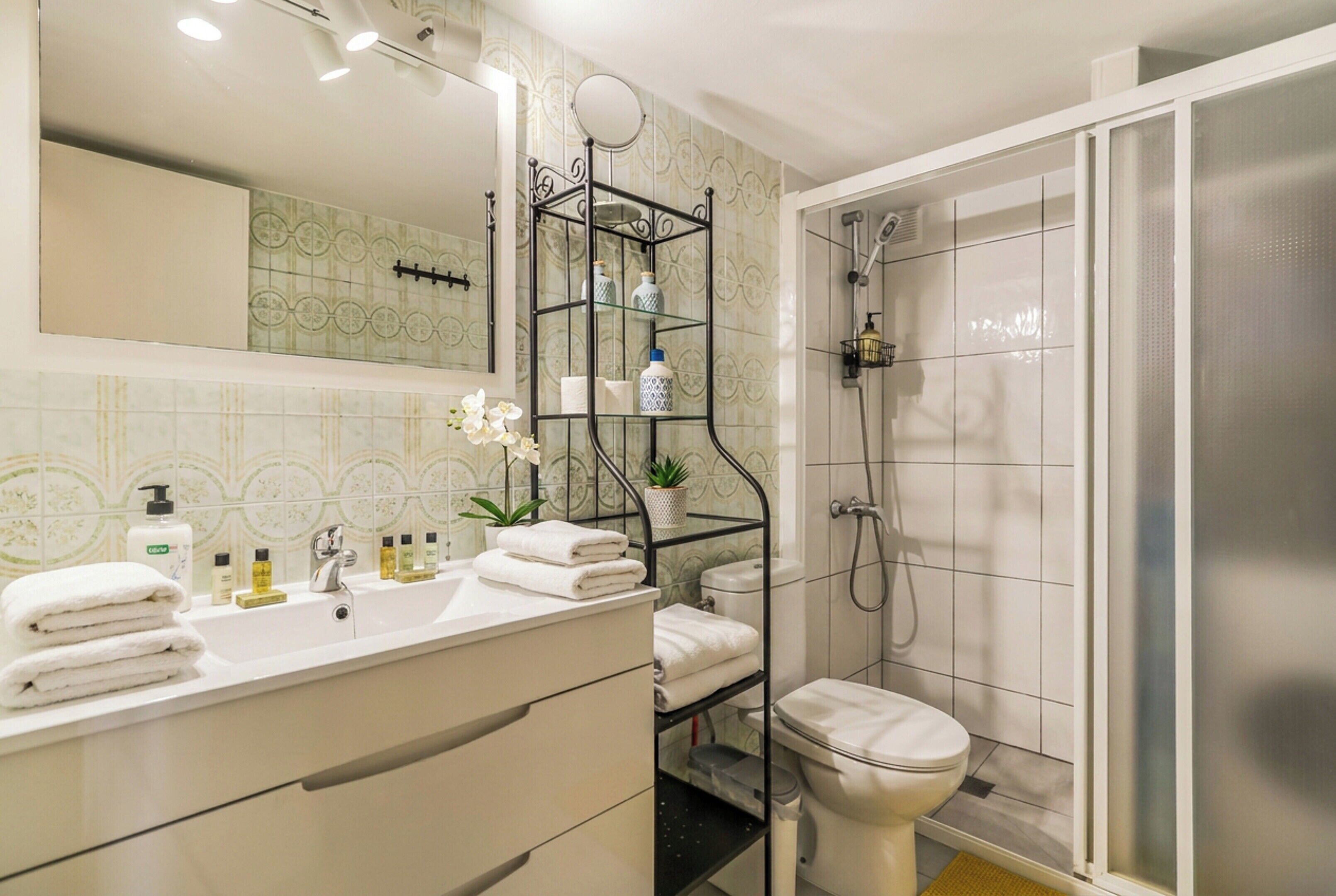 Un baño con lavamanos blanco, espejo y retrete, zona de ducha y estante con artículos de aseo.