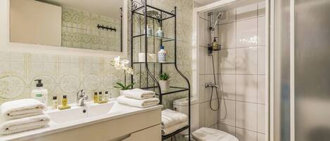 Un baño con lavamanos blanco, espejo y retrete, zona de ducha y estante con artículos de aseo.