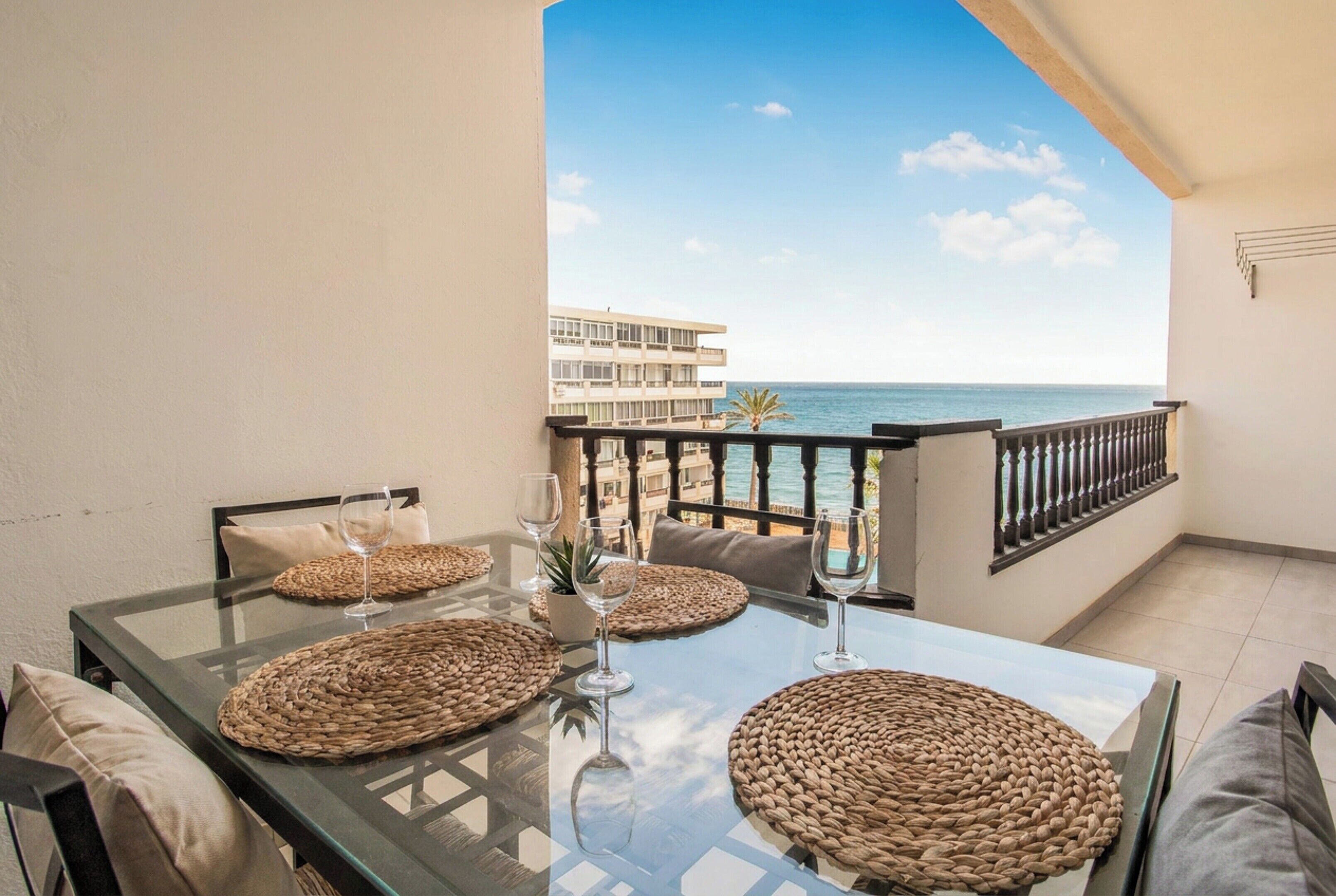A balcony with a glass table set for dining, featuring woven placemats and wine glasses.
