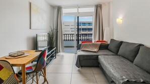 Una sala de estar moderna con un sofá gris, una mesa de comedor de madera y un televisor. Se ve un balcón con barandilla y un edificio al fondo a través de la puerta abierta.