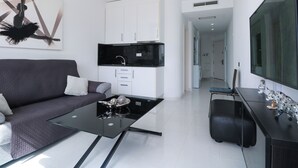A modern living room with a sofa, a glass coffee table, a kitchen area with white cabinets, and a large flat-screen TV. - Cura Marina Seafront Playa del Cura (Mogán)