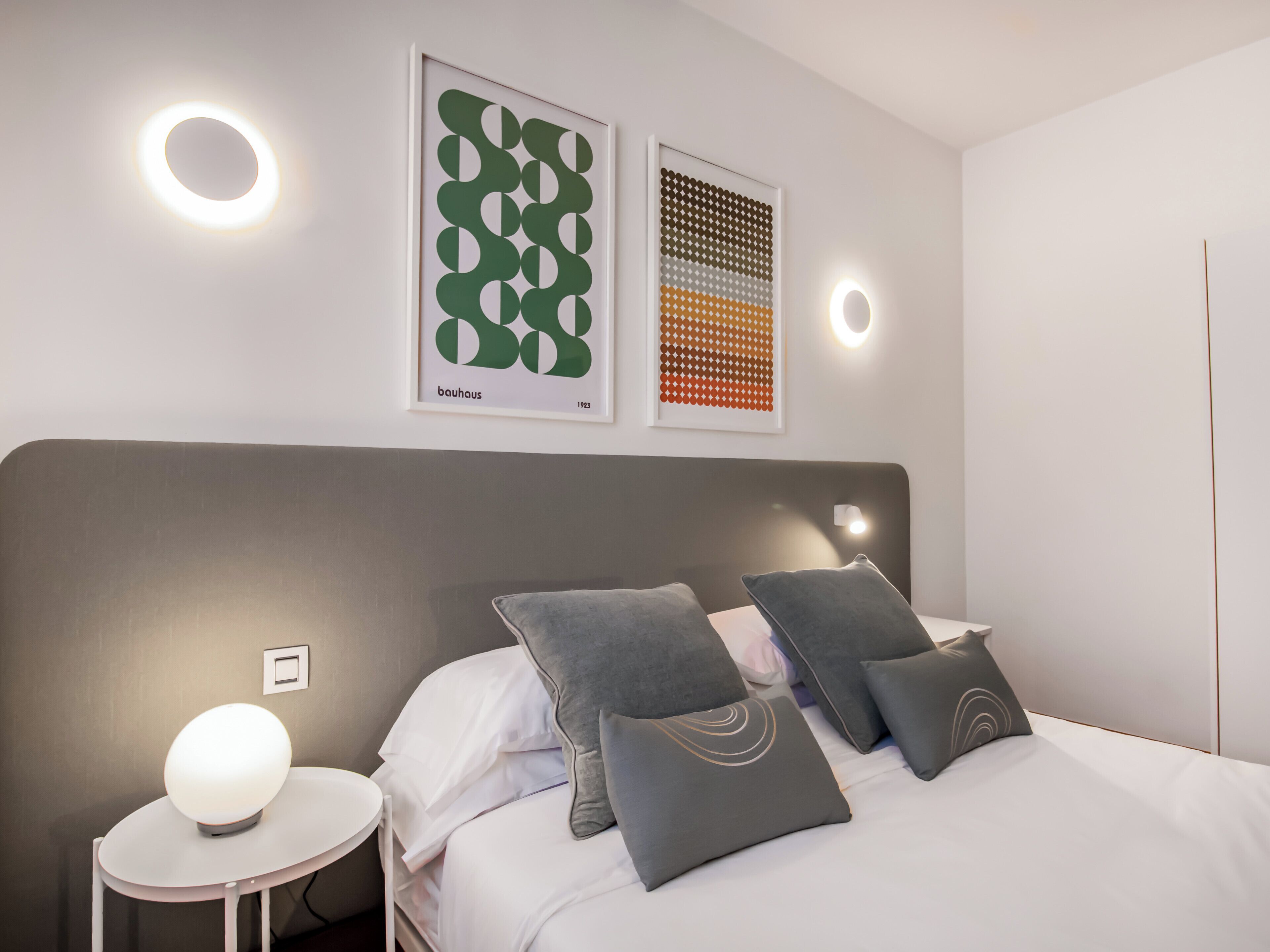 A modern bedroom with a grey headboard, white bedding, and grey pillows. There are two framed artworks on the wall, a round light on the nightstand, and a small table with a white lamp.