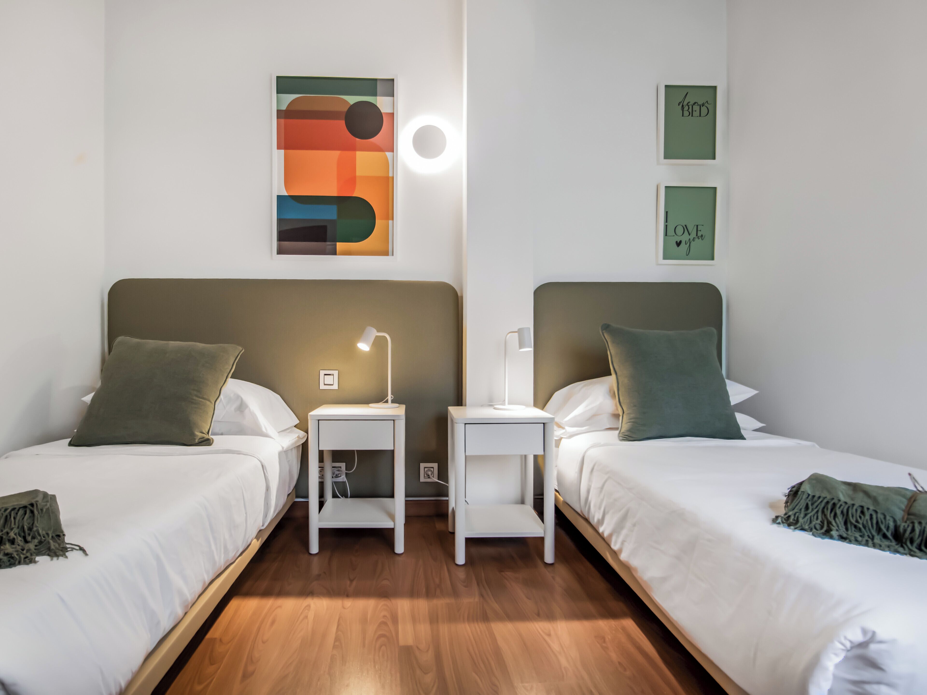 A hotel room with two beds, each with a pillow and a bedside table with a lamp. There are wall art pieces and a light fixture above the beds.