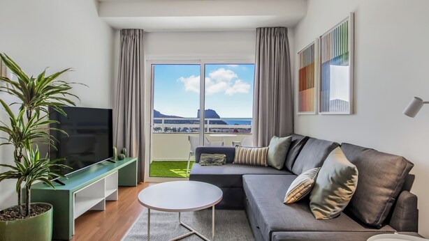 A modern living room with a grey sofa, a small white table, a TV, and a potted plant. - Agaete Shine Suites 302 (Agaete)
