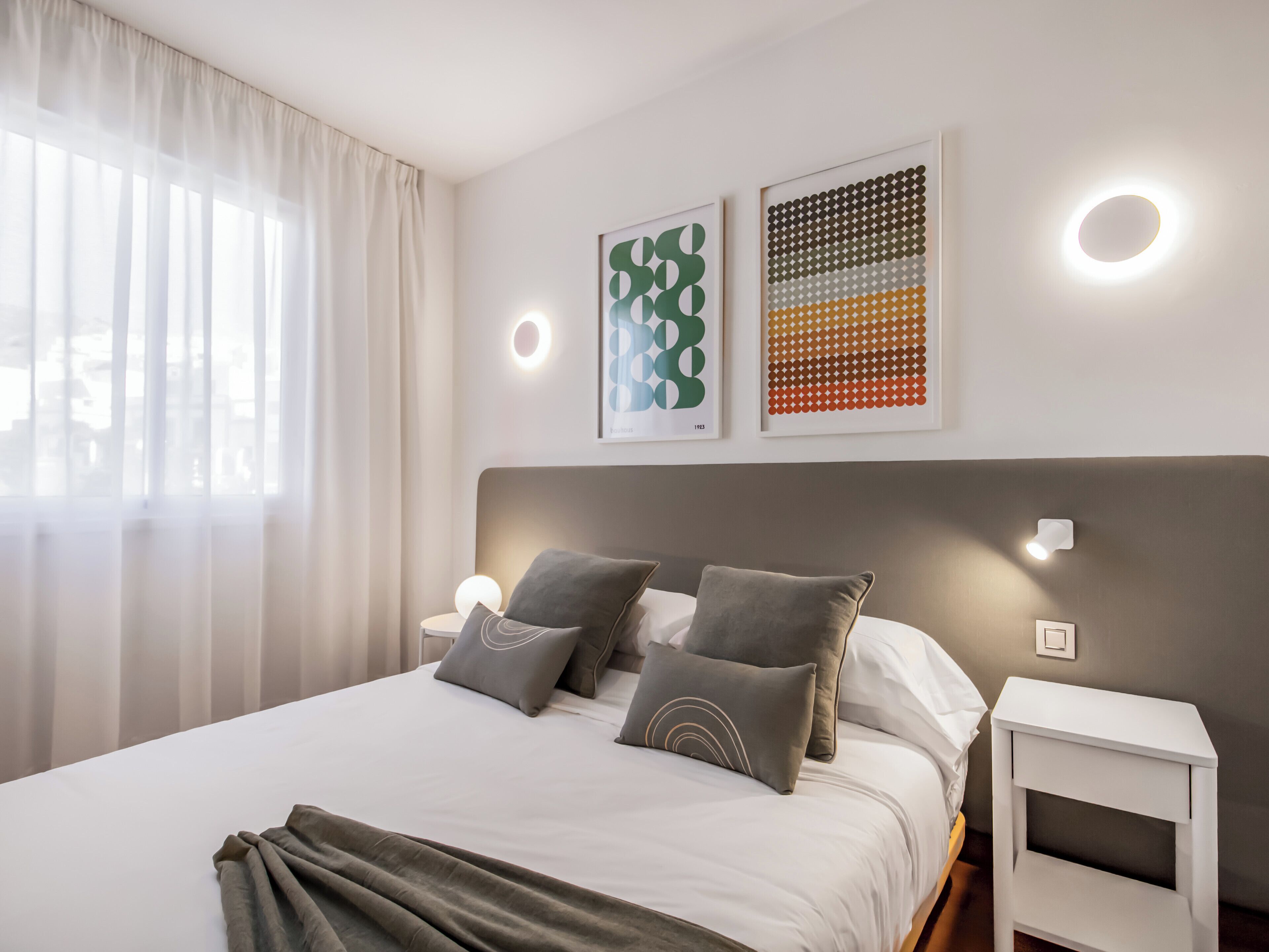 Una cama bien hecha con un cabecero gris, dos mesitas de noche con lámparas, y dos obras de arte abstracto en la pared.