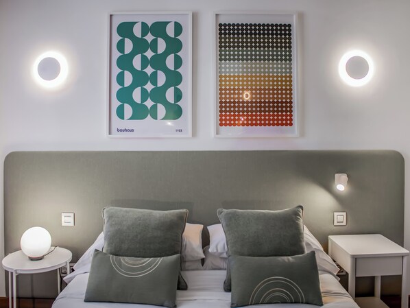 A hotel room with a modern design, featuring a large bed with two pillows, a bedside table with a lamp, and two framed artworks on the wall.