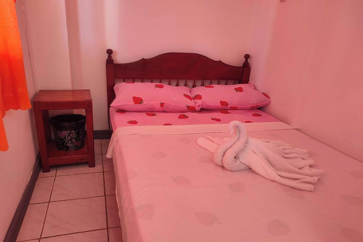 A small, neatly made bed with pink bedding and strawberry-patterned pillows, a wooden nightstand with a trash bin, and a white towel folded on the bed.