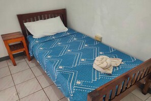 A single bed with a patterned blue bedspread and a white pillow. A wooden nightstand with a lamp is to the left. - Istroberi Inn By Zuzu (Puerto Princesa)