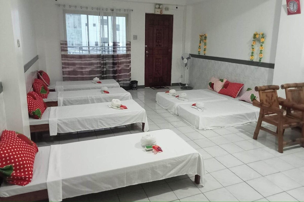 A hotel room with four single beds, each with white bedding and red pillows. There is a wooden chair and a small table with a fan.